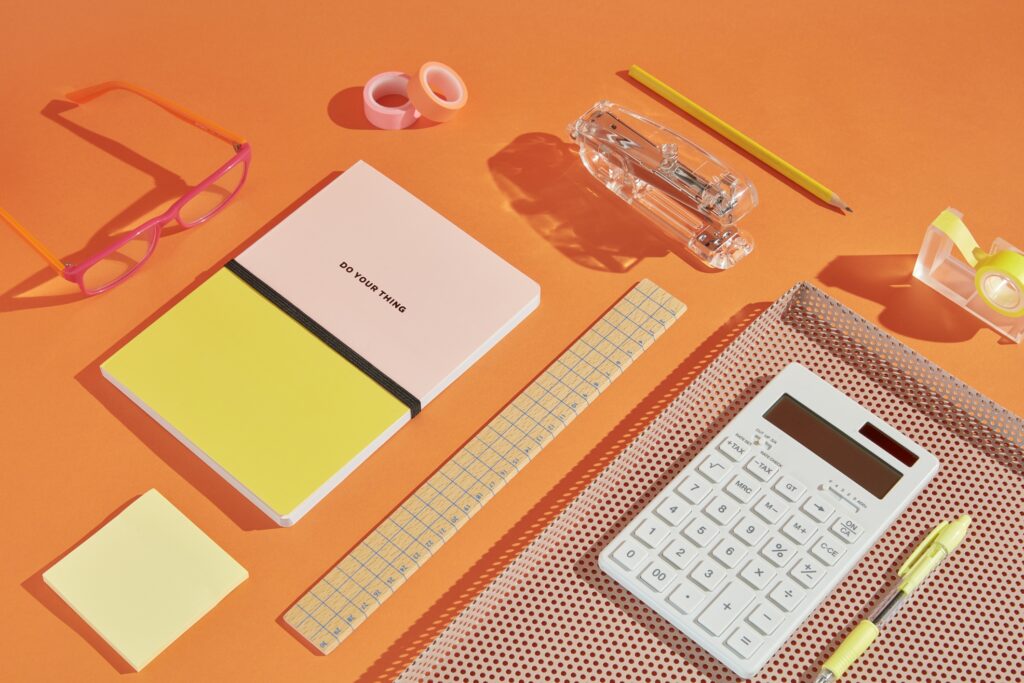 Securinvest editorial still-life flat-lay on an orange backdrop — notebook, ruler, pencil, white calculator, pink glasses and sticky notes, brand-color storytelling set.